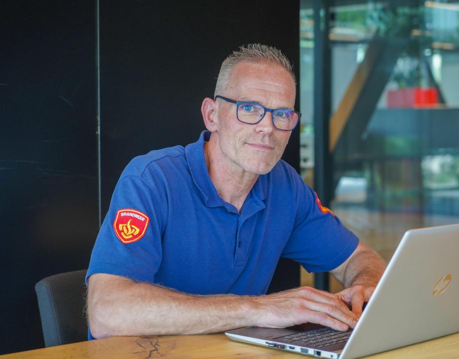 Er zit een man voor een laptop op een kantoor met een polo aan met daarop het embleem van de brandweer.