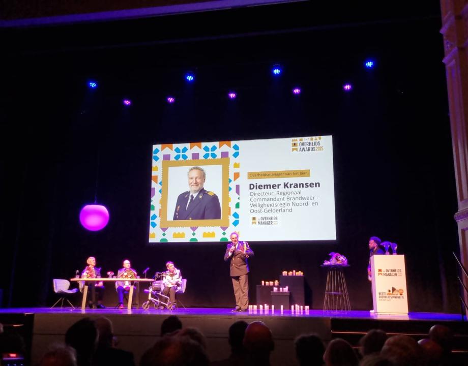 Diemer Kransen staat op de foto op het diascherm. Hij is de winnaar Overheidsmanager van het Jaar geworden. Voor het diascherm waar de afbeelding van Diemer op staat zitten mensen achter een tafel. Dit is de jury.