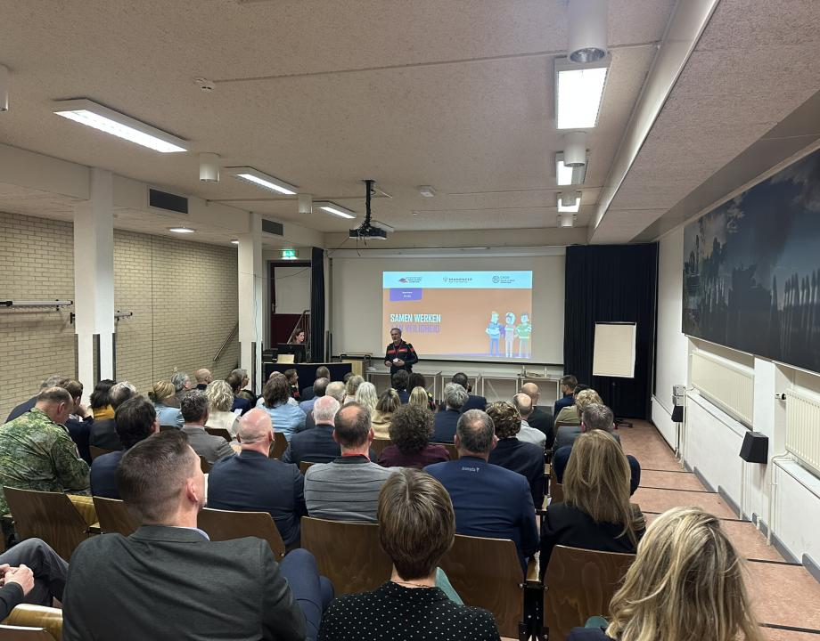 Groep genodigden die naar een presentatie kijken. Een man staat voor het diascherm waar een presentatie op staat