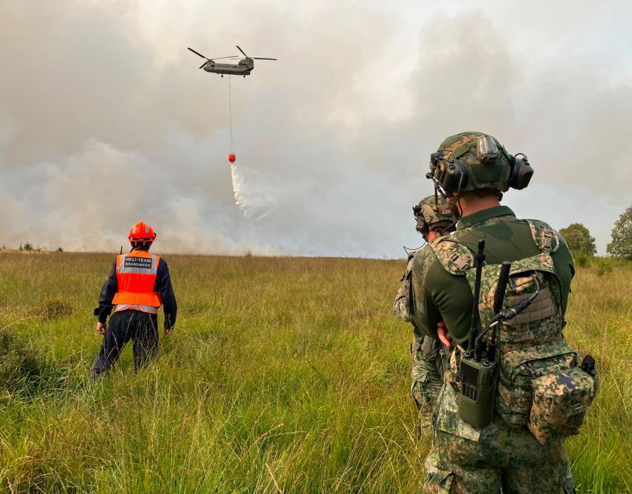 Militaire helikoper lost water vanuit een bucket op natuurbrand, militairen en brandweer kijken toe
