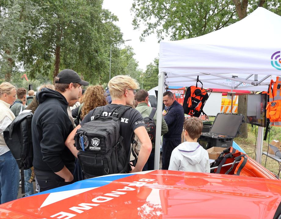 Mensen staan voor een brandweerauto en kijken naar een demo die onder een partytent gehouden wordt